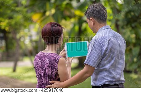 Asian Middle-aged Couple Talking And Look At Tablet Together In Backyard.