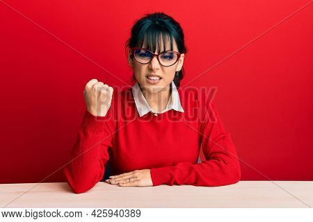 Young brunette woman with bangs wearing glasses sitting on the table angry and mad raising fist frustrated and furious while shouting with anger. rage and aggressive concept. 