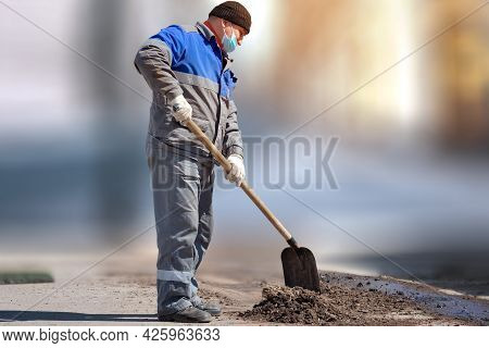 A Laborer In A Medical Mask And Work Clothes Stands With A Shovel On A Blurry Background. Physical L
