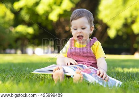 Little Girl, Child Is Studying A Book. Early Toddler Development, Delight Baby Outdoors In The Park.