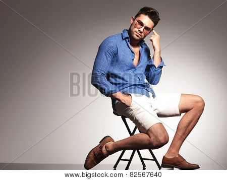 Handsome young fashion man pointing to his head while sitting on a chiar with his hand in pocket.