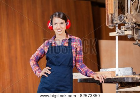 Portrait of confident female carpenter with hand on hip standing by bandsaw in workshop