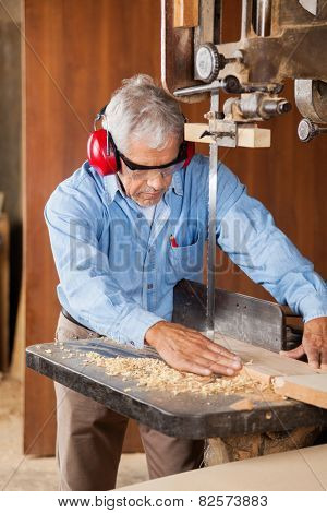 Senior carpenter using bandsaw to cut wooden plank in workshop