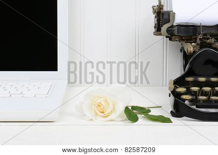 Closeup of a white desk with a rose laying between a modern laptop computer and an antique typewriter. Only half of the laptop and typewriter are shown. Horizontal format.