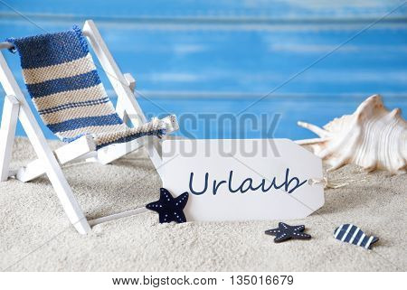 Summer Label With German Text Urlaub Means Holiday Or Vacation. Blue Wooden Background. Card With Holiday Greetings. Beach Vacation Symbolized By Sand, Deck Chair And Shell.