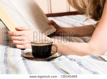 beautiful woman reading a thick book lying on the bed, close-up coffee cup.