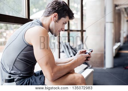 Handsome young portsman listening to music and using his cellphone sitting on windowcill at the gym