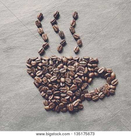 Cup from coffee beans with smoke. Heap of roasted coffee bean on grey stone surface texture. Coffee shop or cafe background. Natural stone and cup from beans. Soft color toning, top view