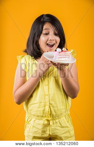 portrait of Indian kid eating cake or pastry, cute little girl eating cake, girl eating strawberry cake or pastry over yellow background