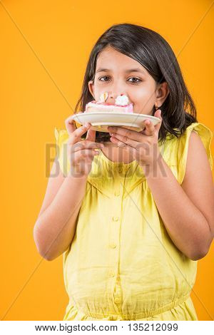 portrait of Indian kid eating cake or pastry, cute little girl eating cake, girl eating strawberry cake or pastry over yellow background