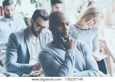 Overthinking info. Handsome African man holding hand on chin and looking thoughtful while sitting on conference with other people