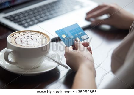 Young beautiful female sitting in modern urban cafe drinking cappuccino and paying with credit card for online shopping on laptop. Back view. Close-up of hands