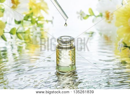 The bottle of Moisturizing cosmetic oil in the water waves on the flowers blur background and pipette with oil drop above the bottle