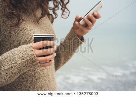Woman With Curly Hair In A Brown Sweater Walks On The Beach, Using Cell Phone And Drinking Coffee