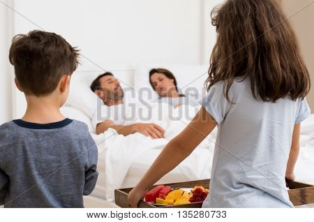 Rear view of daughter and son carrying breakfast for parents in their bed. Parents sleeping peacefully while daughter and son bringing fruits for breakfast.
