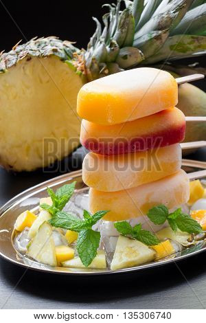 Column of sorbet ice cream popsicles from pineapple mango strawberry on silver plate with ice cubes and fruit pieces on a black background. Pineapple mango strawberry sorbet ice cream popsicles.