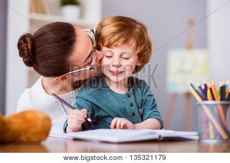 My precious. Young woman with glasses kissing her son while he drawing and sitting at the table