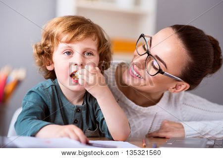 One more piace. Little cute calm boy eating a piece of cake while his smiling beautiful mother looking at him