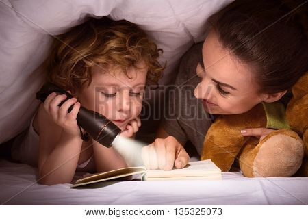 I like my mum. Lovely young mother reading a book to her tired little son after making a hut of blankets on the bed