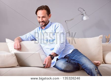 Relaxed time. Relaxed middle-aged man sitting on the couch and using a laptop
