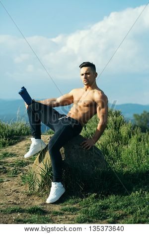 Young sexy man with muscular sexy body and bare torso with chest sitting outdoor with water bottle on sky natural background