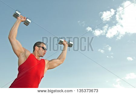 a young man trains with dumbbells outdoors in the red shirt and black shorts