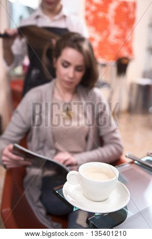 Closeup picture of cup of coffee. Beautiful lady reading magazine while having her hair dobe by hairdresser in hairdressing salon.