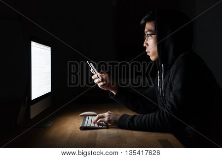 Serious young hacker using laptop and mobile phone in dark room