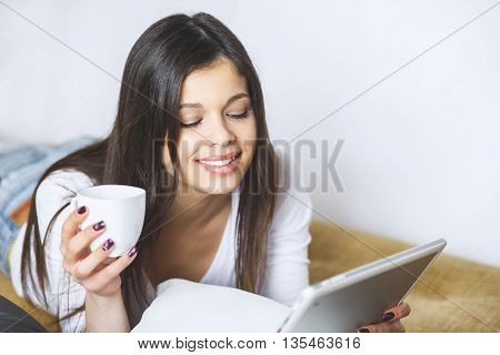 Enjoying her new digital tablet. Beautiful young woman holding touch pad and looking at it with smile. Woman at home relaxing on sofa couch reading email on the tablet computer