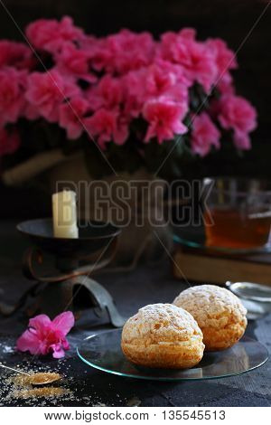 Two custard pastries stuffed with cream on glass saucer on a dark background in the background - azalea flowers