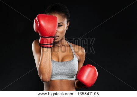 Strong intense young fitness woman covered her face with boxing gloves over black background