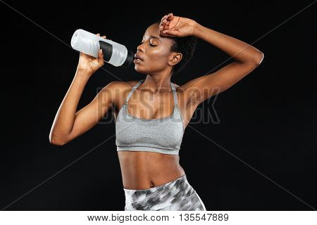 Cute young african american sportswoman standing and holding bottle of water over black background