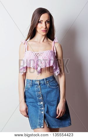 Beautiful young Caucasian woman in pink crochet crop top and denim buttoned skirt posing, smiling. Studio portrait of gorgeous female model in summer outfit. Mild retouch, no filter, studio lighting.