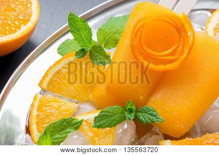 Orange sorbet ice cream decorated with a flower of orange peel mint leaves on silver plate with ice cubes orange slices on black background. Orange sorbet ice cream popsicles. Horizontal. Close.