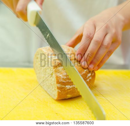 Cutting slices from loaf of bread sitting on yellow surface, hand holding large knife.