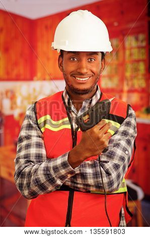 Young engineer carpenter wearing square pattern flanel shirt with red safety vest, holding glue gun smiling to camera.