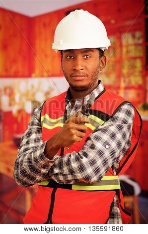Young engineer carpenter wearing helmet, square pattern flanel shirt with red safety vest, holding small handheld electric polisher tool smiling to camera.