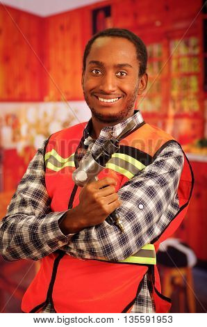 Young engineer carpenter wearing square pattern flanel shirt with red safety vest, holding handheld electric power tool smiling to camera.