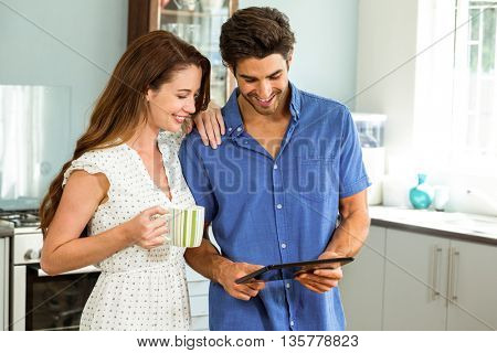 young couple holding cup of coffee while using digital tablet in kitchen