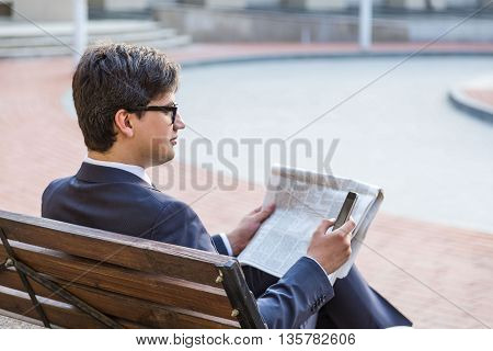 Young attractive businessman sitting on outdoor bench holding news paper and using smart phone