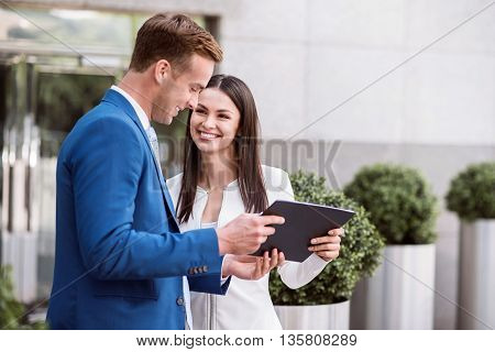 Discuss business affairs. Pleasant cheerful smiling colleagues standing near office building and holding tablet while talking