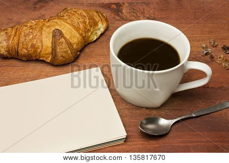 A photo of cup of dark coffee with some cane sugar a spoon a croissant and a blank notepad with copyspace on a dark wooden board texture