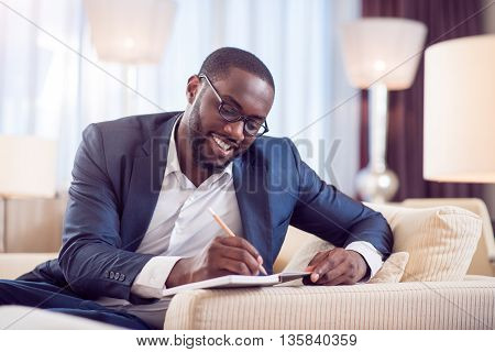 Lets work. Satisfied young afro American man in suit while taking notes in a diary while sitting on the sofa in a hotel