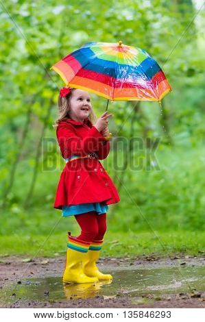 Little girl playing in rainy summer park. Child with colorful rainbow umbrella waterproof coat and boots jumping in puddle in the rain. Kid walking in autumn shower. Outdoor fun by any weather
