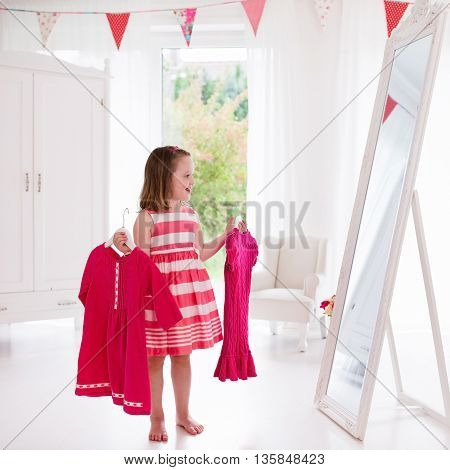 Little girl choosing dresses in white bedroom. Child watching mirror reflection holding pink dress choosing outfit. Girls nursery. Shopping clothing for kids. Dressing room interior for children.