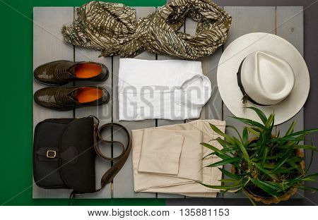 Women's outfit on wooden background. fashion clothes. beige straw hat beige pant white shirt green scarf black crossbody bag and oxford shoes near flower pot. Top view.