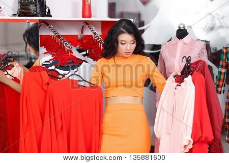 Young woman shopping in a clothing store. Woman shopping for dress in clothing retail store. Caucasian shopper girl choosing red dress in shop during sale. Woman shopping for dress. Fashion shopping