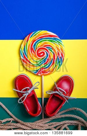 Small red boat shoes near big multi-colored lollipop and rope on colored background. Top view. Close up.