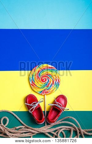 Small red boat shoes near big multi-colored lollipop and rope on colored background. Top view. Copy space frame