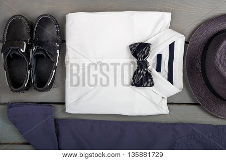 Men's outfit on wooden background. Kids fashion clothes. Grey fedora navy pant white shirt black bow tie and boat shoes for boy. Top view.